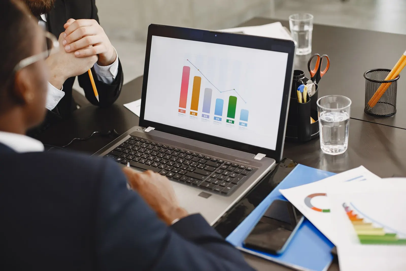 Laptop showing colorful bar charts with a person taking notes beside printed charts and a glass of water