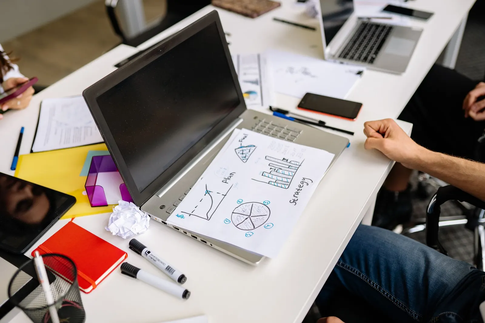 Hand-drawn chart sketches on paper next to a laptop, markers and sticky notes on a desk