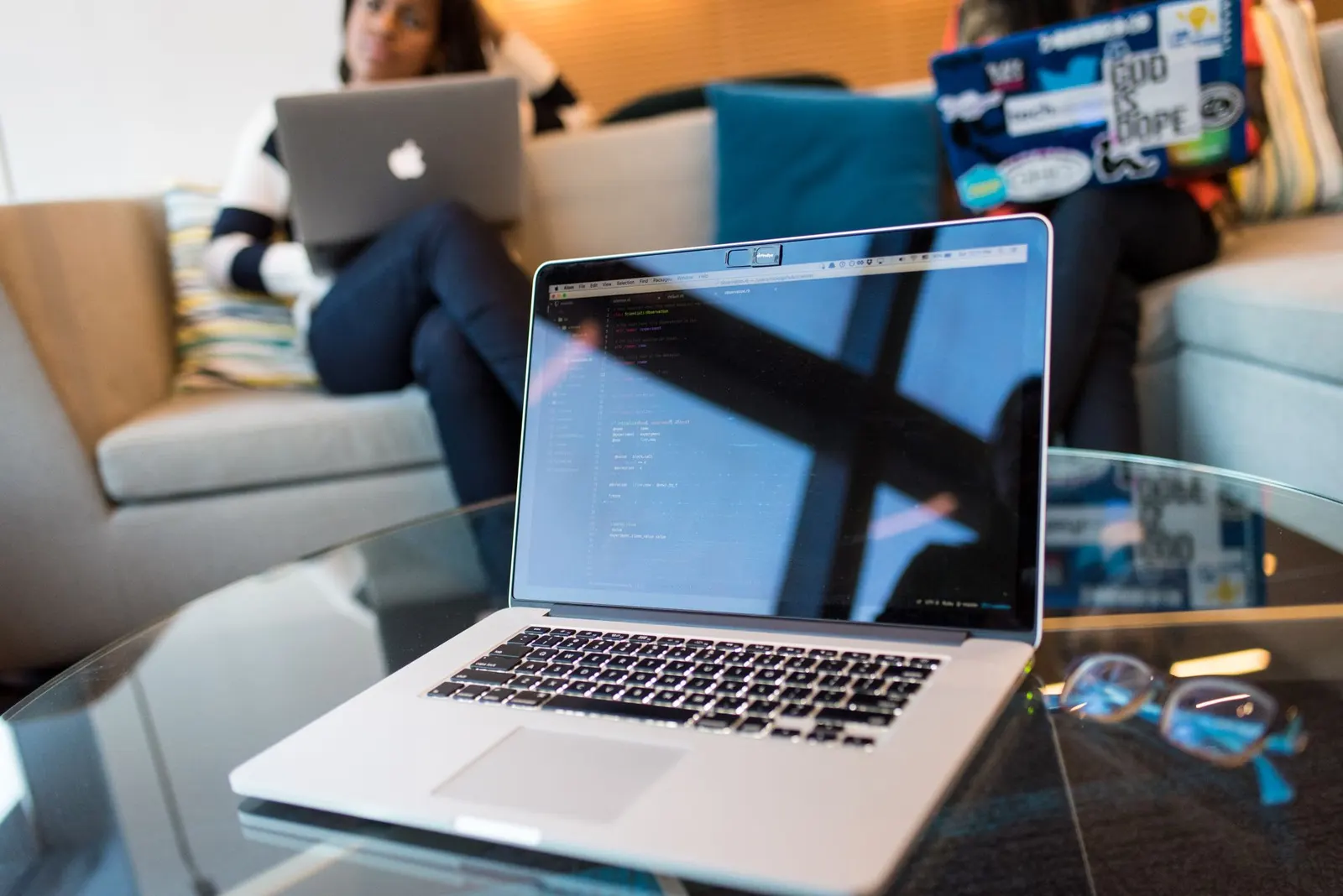 Open laptop with code on screen on a glass table in a bright lounge with people working in the background