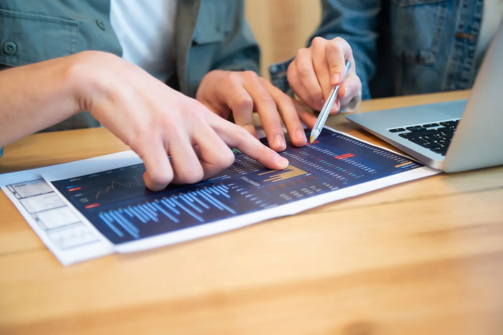 Hands pointing at a tablet displaying blue analytics charts on a wooden desk beside a laptop