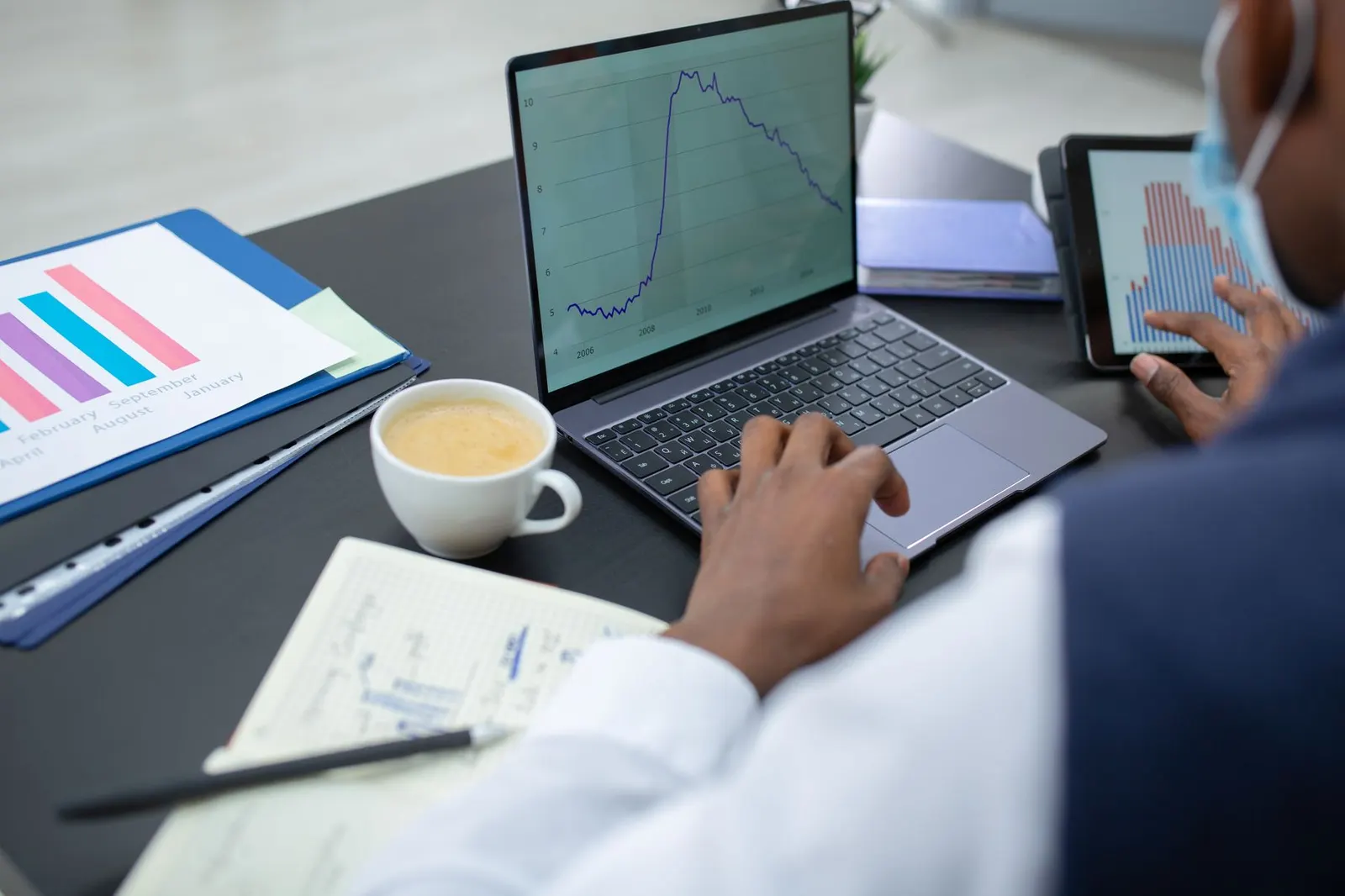 Person working on a laptop showing a line chart with printed reports and a coffee cup on the desk