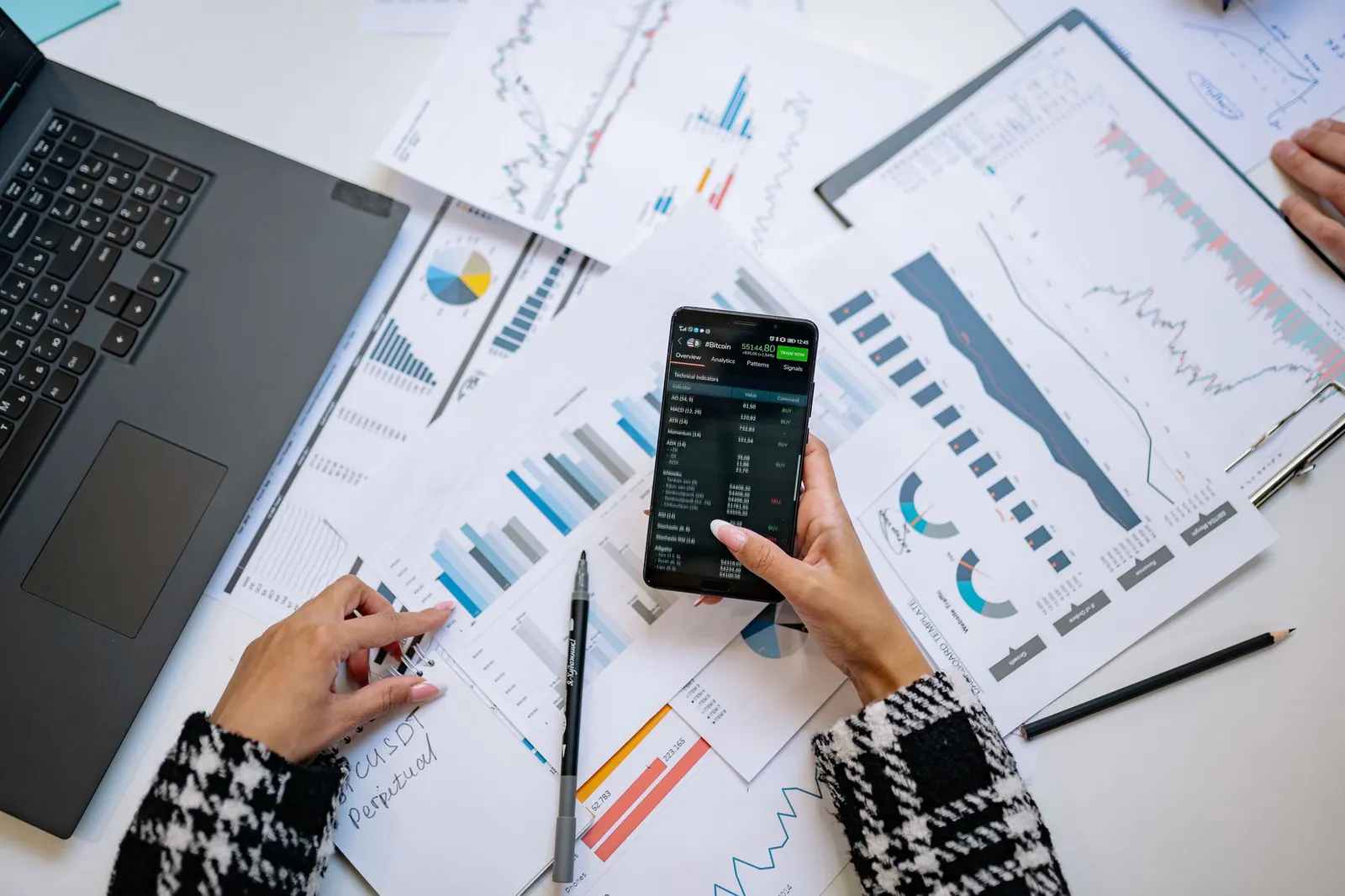 Person holding a smartphone over printed charts and reports spread across a desk next to a laptop