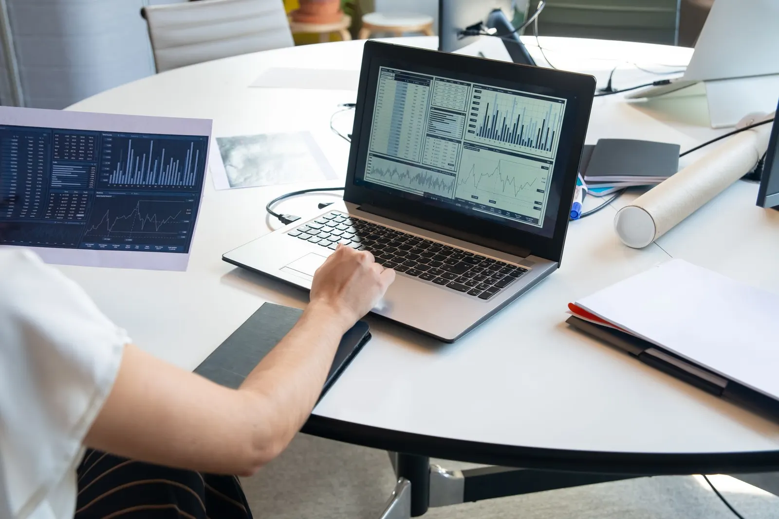 Laptop showing analytics charts on a round office table beside a second monitor with data tables